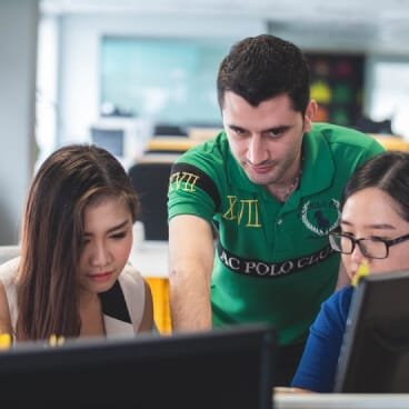 A professional indoor photograph showing a diverse group of young students and a male instructor collaborating in a modern study center or computer lab. A mentor wearing a green polo shirt leans over to guide a female student with glasses working on a computer screen, while another female student looks closely at her monitor. This imagery represents the focused, flexible preparation environment provided by a non attending school in Delhi for students maximizing their entrance prep while retaining the 85% state quota of Delhi benefits for competitive examinations like the Delhi quota in NEET and Delhi quota in JEE.