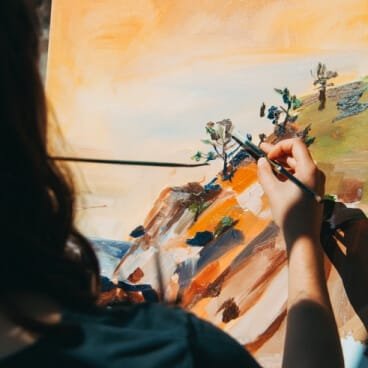 An over-the-shoulder view of a person painting an impressionistic landscape scene onto a canvas with a paintbrush. The painting features rocky terrain, small green trees, and warm orange and yellow tones in the background. This artistic visual serves as a metaphor for the freedom and custom time styling available to students who choose a non attending school in Delhi. By utilizing a flying admission in Delhi, students can pursue personal interests while maintaining full eligibility for competitive advantages like the Delhi state quota benefit in NEET and the standard Delhi quota in JEE.