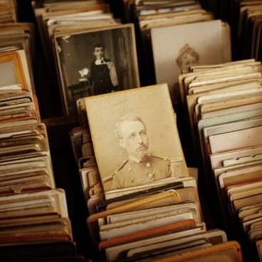 A close-up, warm-toned photograph showing stacks and rows of vintage, sepia-toned portraits and antique photographs organized in an archival box or filing drawer. In the center, a clear portrait shows a historical military figure with a mustache in uniform, surrounded by other old photographs of people from a past era. This archival image serves as a visual metaphor for organizing records and securing credentials when enrolling in a non attending school in Delhi. It represents the careful registration needed for a flying admission in Delhi to safely access high-value institutional benefits like the 85% Delhi state quota in NEET and the powerful Delhi quota in JEE