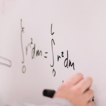 A close-up photograph showing a person's hand writing mathematical integration formulas with a black marker on a white marker board. The equations displayed include calculus integral signs with variables r squared dm integrated from limits zero to L. This academic visual represents the rigorous, uninterrupted preparation environment achieved by choosing a non attending school in Delhi. By enrolling in a flying admission in Delhi, science students can invest their day-to-day focus into intensive physics and math problem-solving while safely preserving their high-value eligibility for the 85% Delhi state quota in NEET and the competitive Delhi quota in JEE.